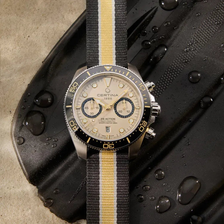 The Certina DS Action Diver Chrono placed on a wet black diving fin, emphasizing its rugged and water-resistant design. The beige dial features gold-tone accents, two black chronograph subdials, and a date window at 6 o’clock. The unidirectional black ceramic bezel with gold markings surrounds the dial, while the stainless steel case is fitted with polished pushers and a screw-down crown. The black, yellow, and white striped textile NATO strap adds a sporty and adventurous touch. Water d