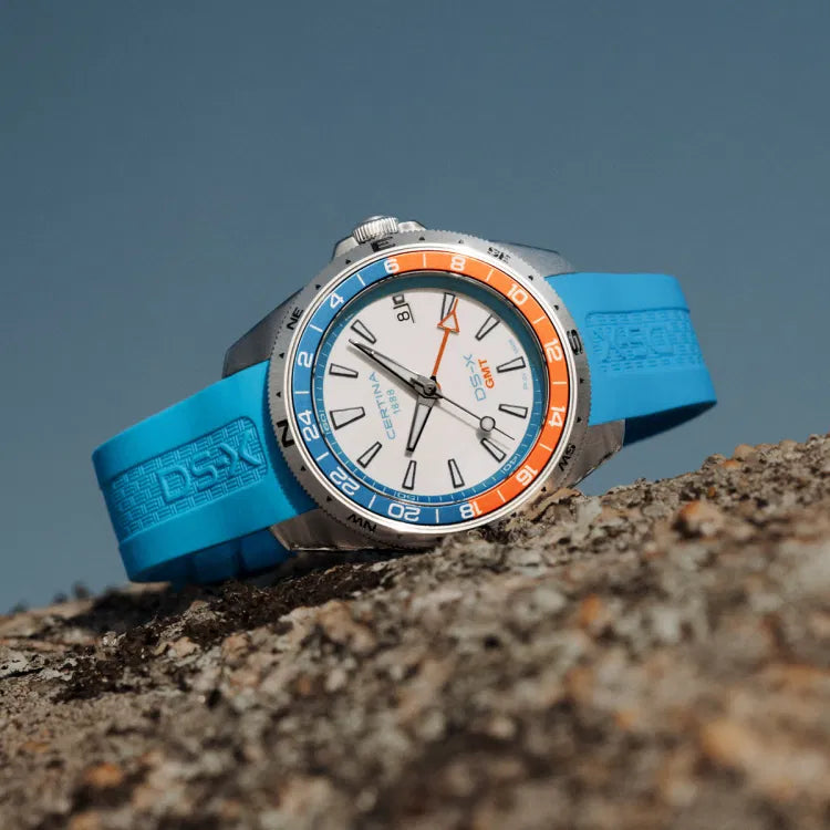 Certina DS-X GMT C047.452.17.011.00 placed on a rock surface, displaying the white dial with GMT hand, orange and blue bezel, and blue rubber strap with DS-X embossing.
