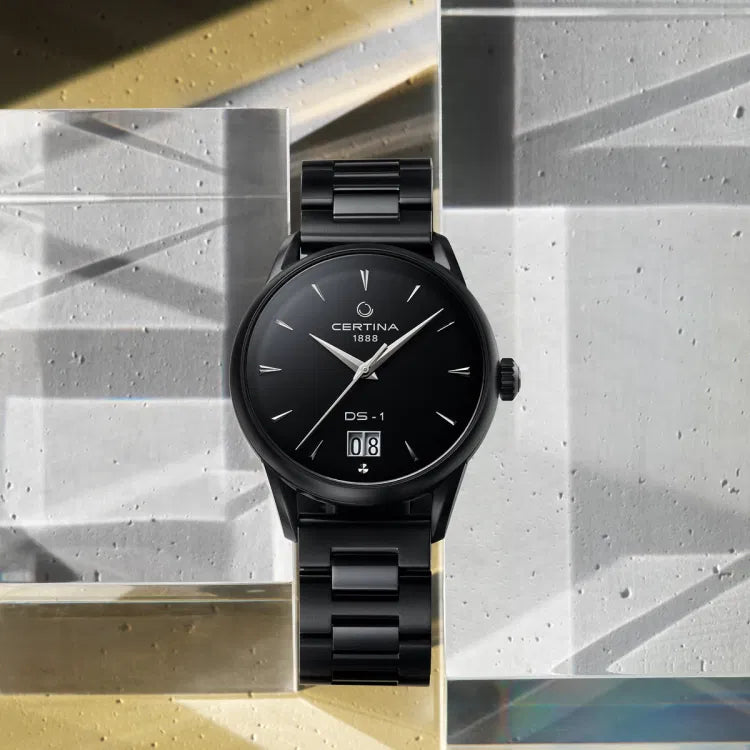 Front view of Certina DS-1 Big Date Powermatic 80 watch with black dial and black PVD-coated stainless steel bracelet, displayed upright on geometric glass and concrete blocks. Dial features ‘CERTINA 1888’, ‘DS-1’, and a large double-digit date window at 6 o’clock.