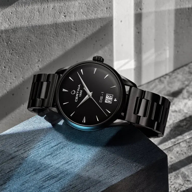Angled view of Certina DS-1 Big Date Powermatic 80 watch resting on a dark blue wooden block. The black dial displays ‘CERTINA 1888’, ‘DS-1’, and a prominent double-digit date window at 6 o’clock. The case and bracelet are black PVD-coated stainless steel.
