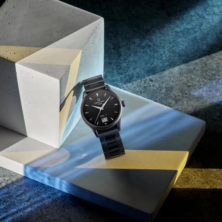 Lifestyle shot of the Certina DS-1 Big Date Powermatic 80 watch with black dial and black PVD-coated stainless steel bracelet, displayed on angled concrete blocks with geometric light and shadow accents. The dial shows ‘CERTINA 1888’, ‘DS-1’, and a large date window at 6 o’clock.