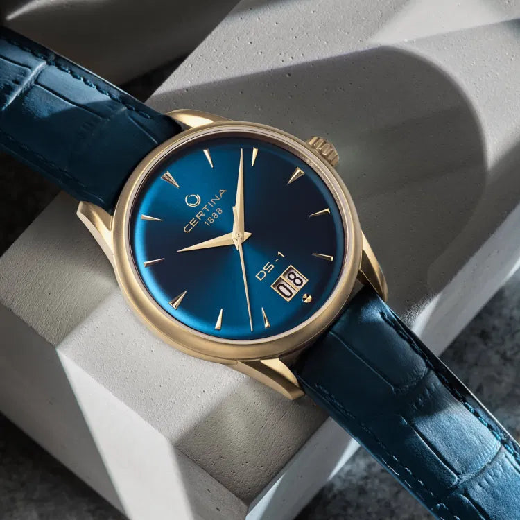 "Certina DS-1 Big Date Powermatic 80 watch with blue dial and gold-tone PVD case, resting diagonally on a textured light gray concrete block. The watch features gold hour markers, a big date display at 6 o’clock, and a blue leather strap."