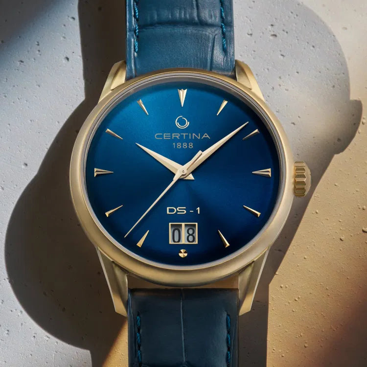 Close-up front view of the Certina DS-1 Big Date Powermatic 80 watch with a blue sunray dial. The dial displays gold-tone hands and markers, the Certina 1888 logo, and “DS-1” above the prominent big date window showing “08”. The watch features a gold-tone PVD-coated stainless steel case and a matching blue leather strap.