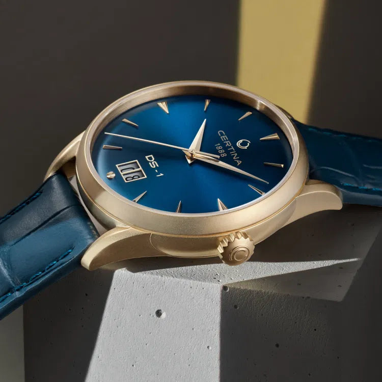 Angled close-up of the Certina DS-1 Big Date Powermatic 80 watch featuring a blue sunray dial with gold-tone hands and indices. The gold-tone PVD-coated stainless steel case includes a signed crown and a prominent big date display at 6 o'clock. The watch is paired with a textured blue leather strap.