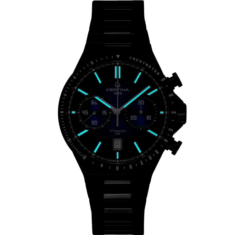 Night view of Certina DS-7 Chrono Auto watch showing glowing blue Super-LumiNova on hands and indices. Chronograph sub-dials and date window at 6 o'clock remain visible on the blue dial. Black PVD-coated titanium case and bracelet.