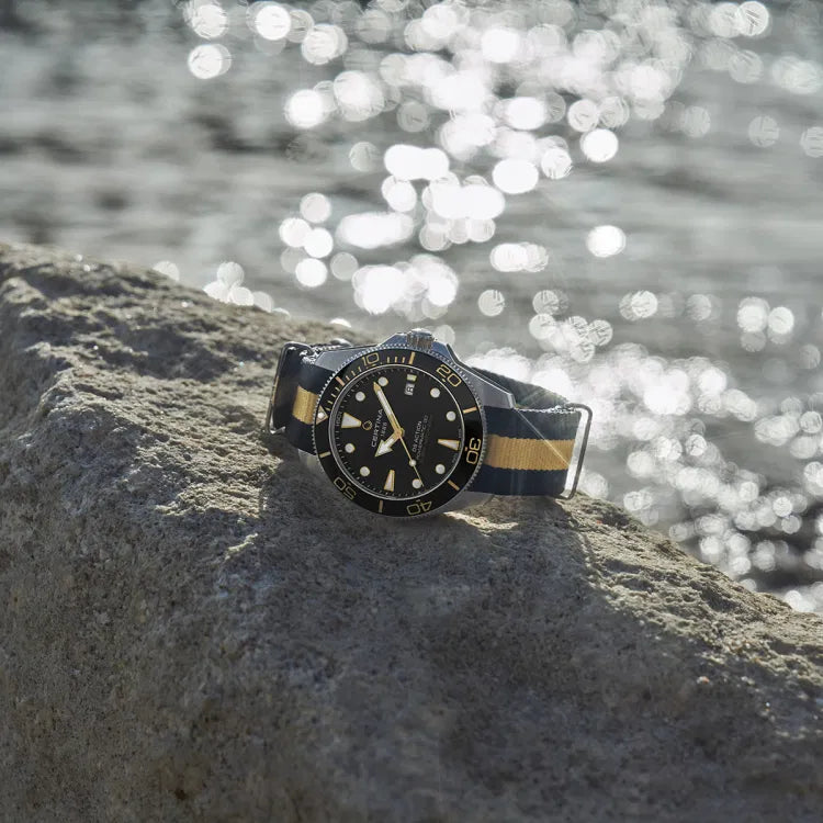 The Certina DS Action Diver 40.5mm watch resting on a sunlit rock by the sea. The watch features a black dial with gold accents, black ceramic bezel, and a black-and-beige NATO strap. Sparkling reflections from the water in the background highlight its marine-ready design.