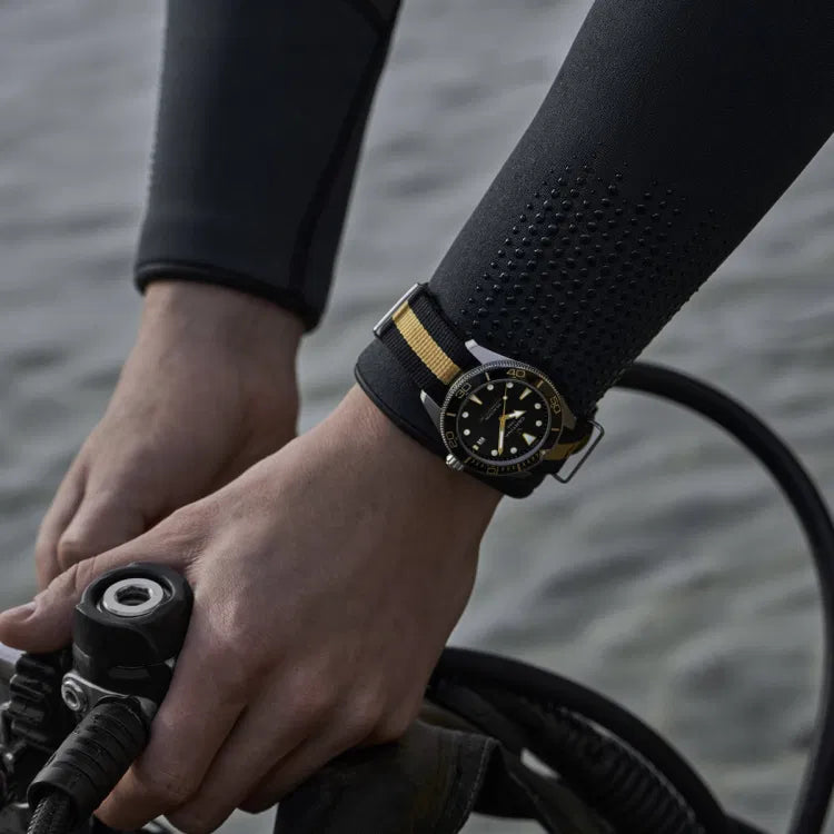Close-up of a diver’s hands in a wetsuit handling scuba equipment, with the Certina DS Action Diver 40.5mm watch clearly visible on the wrist. The watch shows a black dial, gold accents, and a black and beige NATO strap, reinforcing its rugged and aquatic purpose.