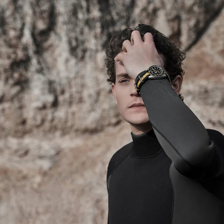 Male model in a black wetsuit standing against a rocky cliff, wearing the Certina DS Action Diver 40.5mm watch with a black and beige NATO strap. The watch features a black dial, gold-toned hands and indices, and a black ceramic bezel with gold markings. The model has his hand raised, partially covering his face, with the watch prominently visible on his wrist.
