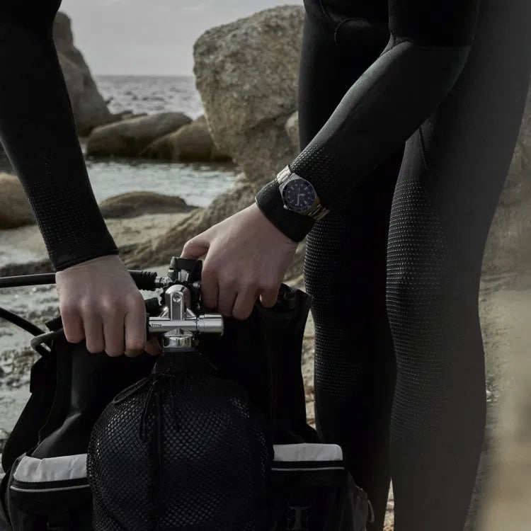 Two divers in wetsuits preparing scuba gear on a rocky shoreline, with one wearing the Certina DS Action Diver 40.5mm titanium watch. The blue dial and ceramic bezel are visible on the wrist, emphasizing the watch’s professional diving design and robust build.