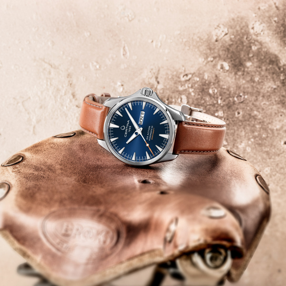 Men's Certina DS Action Day-Date Powermatic 80 watch resting sideways on a leather stool, highlighting the stainless steel case profile, polished finish, and brown leather bracelet. The sleek dial with day and date windows is partially visible, contrasting elegantly with the textured leather surface. A stylish and robust timepiece designed for both everyday wear and adventure