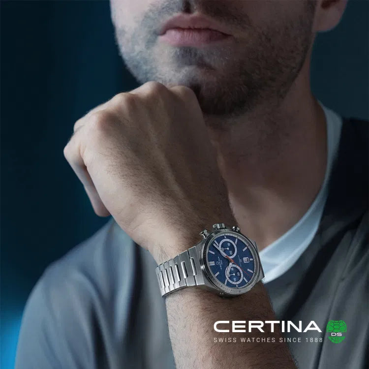 A man wearing the Certina DS-7 Chrono Auto C043.427.11.041.00 on his wrist, with his hand resting near his face in a casual pose. The watch features a deep blue dial with silver-tone indices, chronograph subdials, and a stainless steel bracelet. The lighting emphasizes the watch’s polished stainless steel case and refined details, complementing the modern and sporty aesthetic. The Certina logo and "Swiss Watches Since 1888" branding appear in the lower right corner of the image.