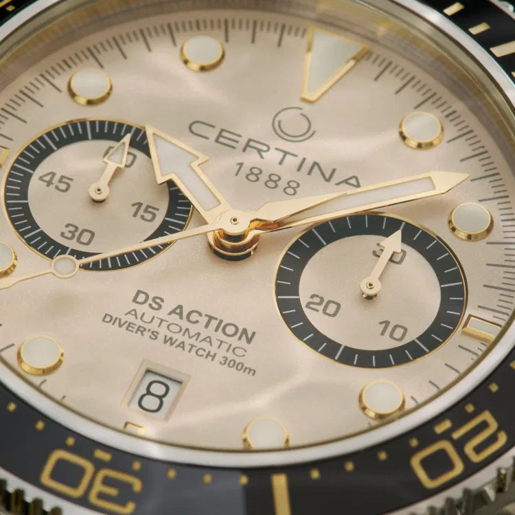 Close-up view of the Certina DS Action Diver Chrono (C032.827.18.271.00) dial, showcasing the intricate details of its beige sunburst finish. The gold-tone hour markers and hands are coated with Super-LumiNova for enhanced readability. The two black subdials provide chronograph functionality, with the left subdial serving as a 30-minute counter and the right displaying running seconds. The black ceramic bezel features gold-tone markings, while the text on the dial includes "Certina 1888" below the 12 o’cloc