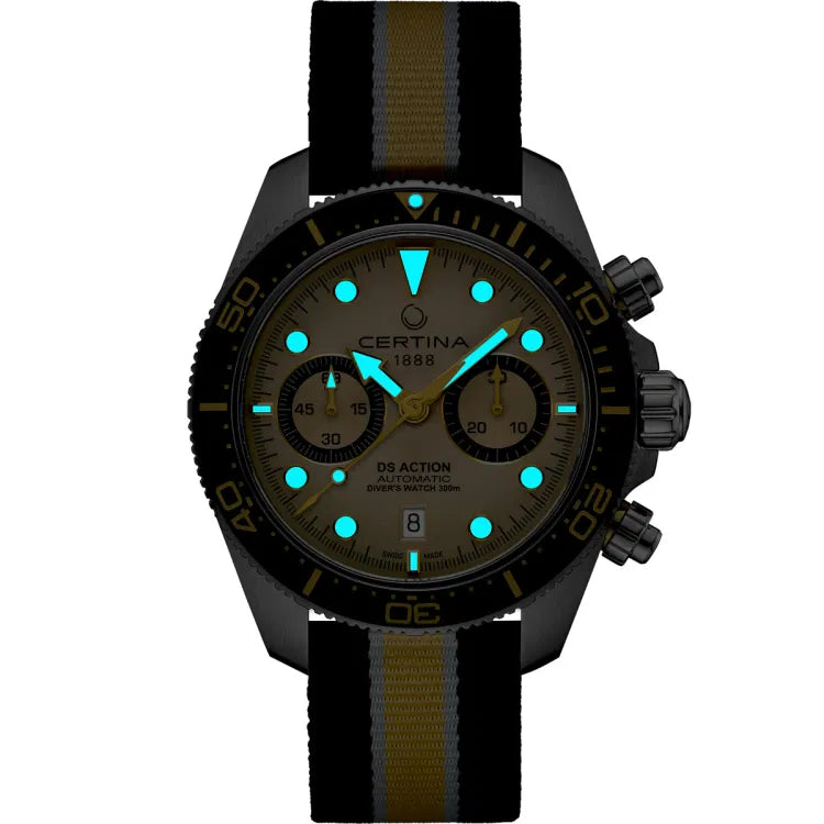 The Certina DS Action Diver Chrono (C032.827.18.271.00) displayed in a dark setting, highlighting its luminescent Super-LumiNova details. The round hour markers, hands, and the triangular bezel marker emit a bright blue-green glow for enhanced readability in low-light conditions. The beige dial, black ceramic bezel with gold-tone markings, and chronograph subdials are subtly visible. The stainless steel case features a screw-down crown and pushers on the right side. The black, yellow, and white striped text