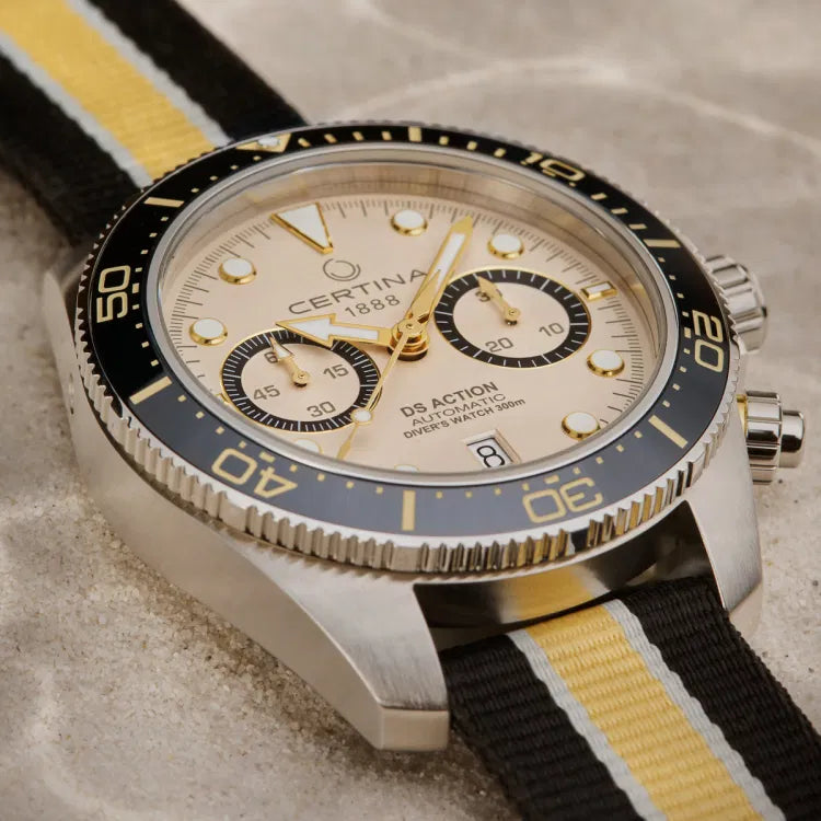 The Certina DS Action Diver Chrono lying on sand, emphasizing its rugged and adventure-ready design. The beige dial features gold-tone accents, two black chronograph subdials, and a date window at 6 o’clock. The unidirectional black ceramic bezel with gold-tone markings provides precise dive timing, while the brushed stainless steel case enhances durability. The screw-down crown and chronograph pushers are polished, reflecting the watch’s refined craftsmanship.