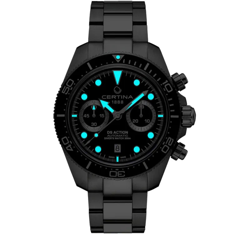 Front view of the Certina DS Action Diver Chrono in a dark setting, highlighting the Super-LumiNova-coated hands and hour markers glowing blue. The black dial features 'Certina 1888' below 12 o’clock and 'DS Action Automatic Diver’s Watch 300m' above 6 o’clock. The ceramic bezel with a 60-minute scale remains visible, along with the chronograph subdials at 9 and 3 o’clock. The stainless steel bracelet and case blend into the dark background, emphasizing the luminous elements.