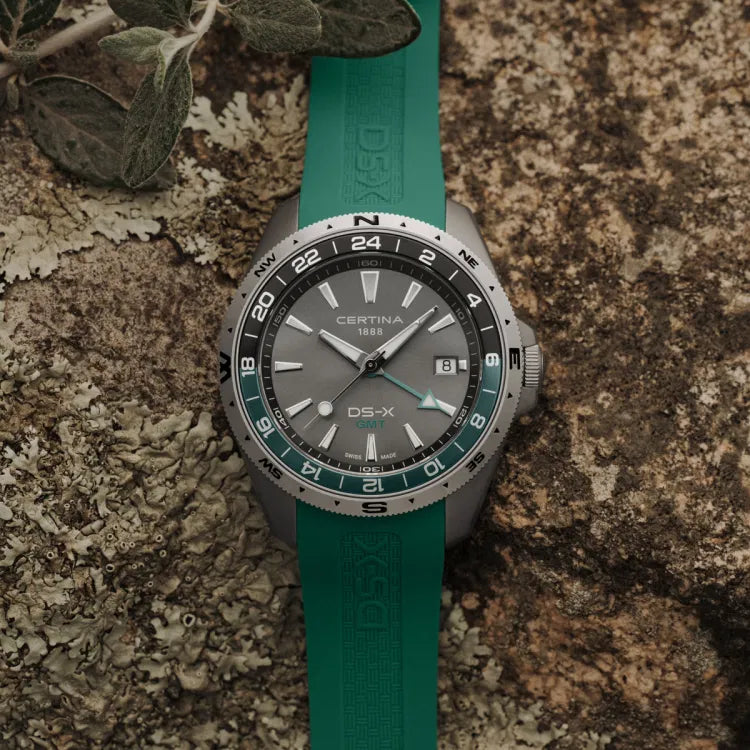certina ds-x gmt c047.452.17.081.00 watch with green rubber strap and green-black gmt bezel, grey dial with certina 1888 and ds-x gmt logos, placed on natural stone surface with leaves