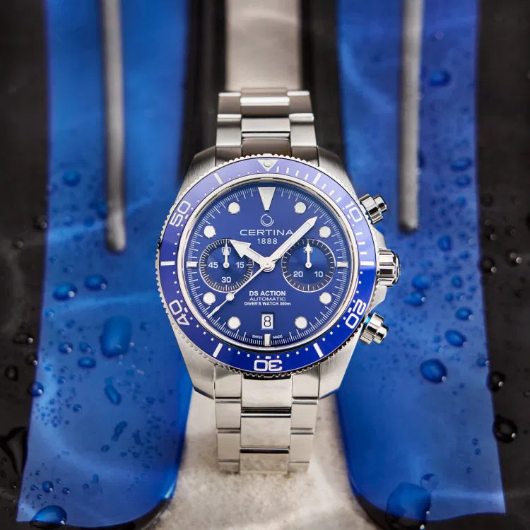 Certina DS Action Diver Chrono with a blue dial placed on wet diving fins, emphasizing its 300m water resistance. The dial features chronograph subdials, Super-LumiNova-coated markers, and a date display at 6 o’clock. The unidirectional ceramic bezel with a 60-minute scale ensures precise timing. The stainless steel case and bracelet contrast with the wet surface, highlighting durability. Water droplets on the fins enhance the watch’s professional diving aesthetic.