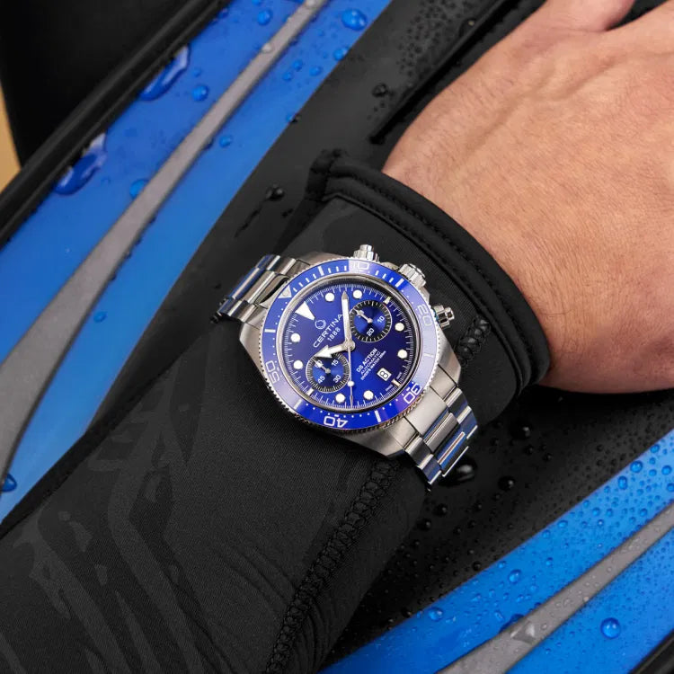 Certina DS Action Diver Chrono worn over a wetsuit sleeve, emphasizing its professional diving design. The blue dial features chronograph subdials, Super-LumiNova-coated markers, and a date display at 6 o’clock. The unidirectional ceramic bezel with a 60-minute scale ensures precise timing. The stainless steel bracelet contrasts with the dark wetsuit, highlighting its rugged yet refined look. Positioned against wet diving fins, the image showcases the watch’s 300m water resistance and suitability for diving