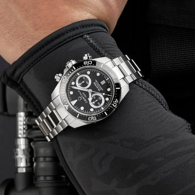 Certina DS Action Diver Chrono worn over a wetsuit sleeve, highlighting its professional diving design. The black dial features chronograph subdials, Super-LumiNova-coated markers, and a date display at 6 o’clock. The unidirectional ceramic bezel with a 60-minute scale ensures precise timing. The stainless steel bracelet contrasts with the dark wetsuit, emphasizing its rugged yet elegant look. Positioned against diving equipment, the image showcases the watch’s 300m water resistance & suitability for diving