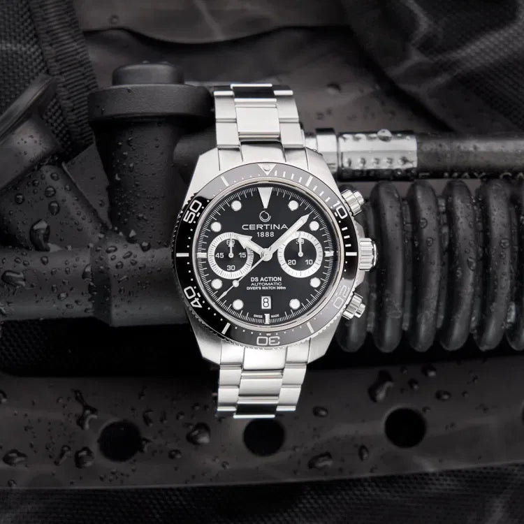 Certina DS Action Diver Chrono placed on wet diving equipment, emphasizing its 300m water resistance. The black dial features chronograph subdials, Super-LumiNova-coated markers, and a date display at 6 o’clock. The stainless steel case and bracelet contrast with the dark, water-drenched background, highlighting the watch’s durability. The unidirectional ceramic bezel with a 60-minute scale ensures precise timing for divers. Water droplets on the surface enhance the rugged, professional diving aesthetic.