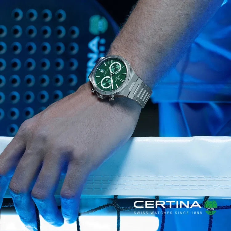 A man wearing the Certina DS-7 Chrono Auto C043.427.11.091.00 with a striking green dial and stainless steel bracelet. His wrist rests on a net, with a blue-lit sports environment in the background, emphasizing the watch’s sporty yet elegant appeal. The chronograph subdials, tachymeter bezel, and luminous hands stand out against the bold green dial. The Certina logo and "Swiss Watches Since 1888" branding appear in the bottom right corner, reinforcing the brand’s heritage and craftsmanship.